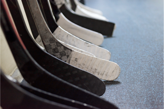 A hockey dressing-room shot focused in on various carbon fiber composite ice hockey sticks displaying various models and various tape jobs featuring either black or white cloth hockey tape