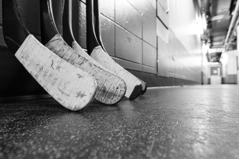 Three in-stock hockey sticks with taped blades and unique hockey stick graphics rest against a wall in a hallway, shown in black and white.