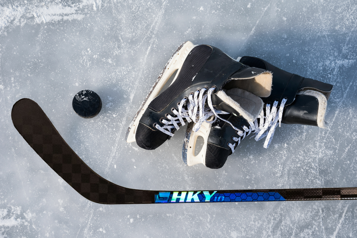 Hockey skates with white laces and a puck lie on an icy surface beside an "HKY" branded stick, conveying a sense of sport and readiness.