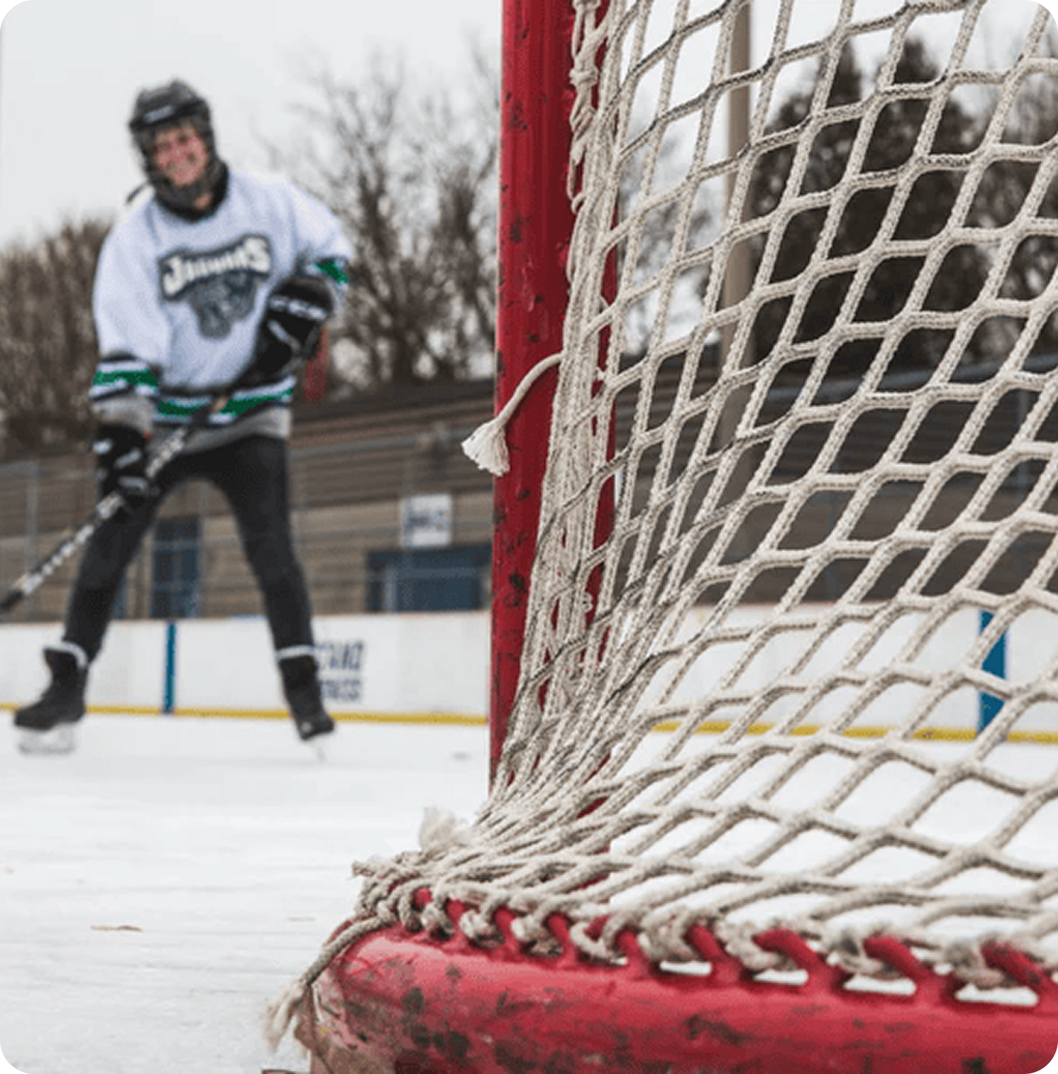Close-up of a red hockey net in focus, with an out-of-focus player in a white jersey skating in the background on an outdoor rink, conveying action.